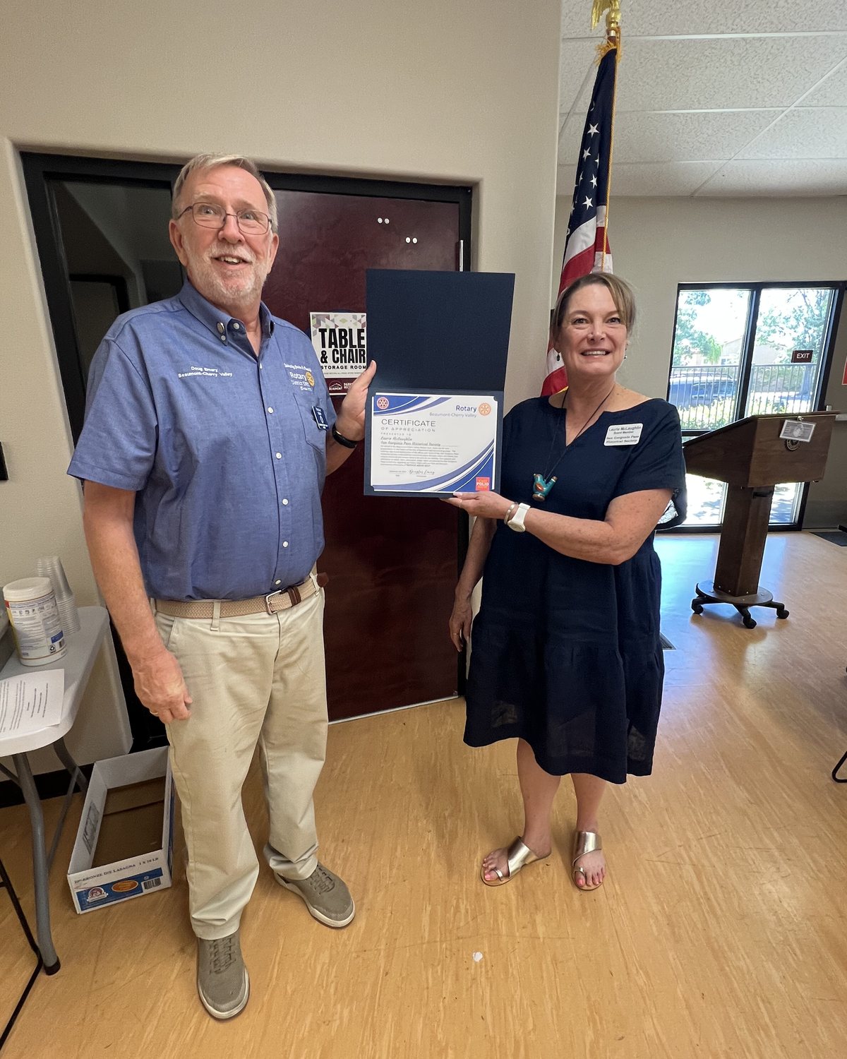 President Doug Emery and Laurie McLaughlin of the San Gorgonio Pass Historical Society hold an open folder containing her Certificate of Appreciation, beside the American flag at the Chatigny Center