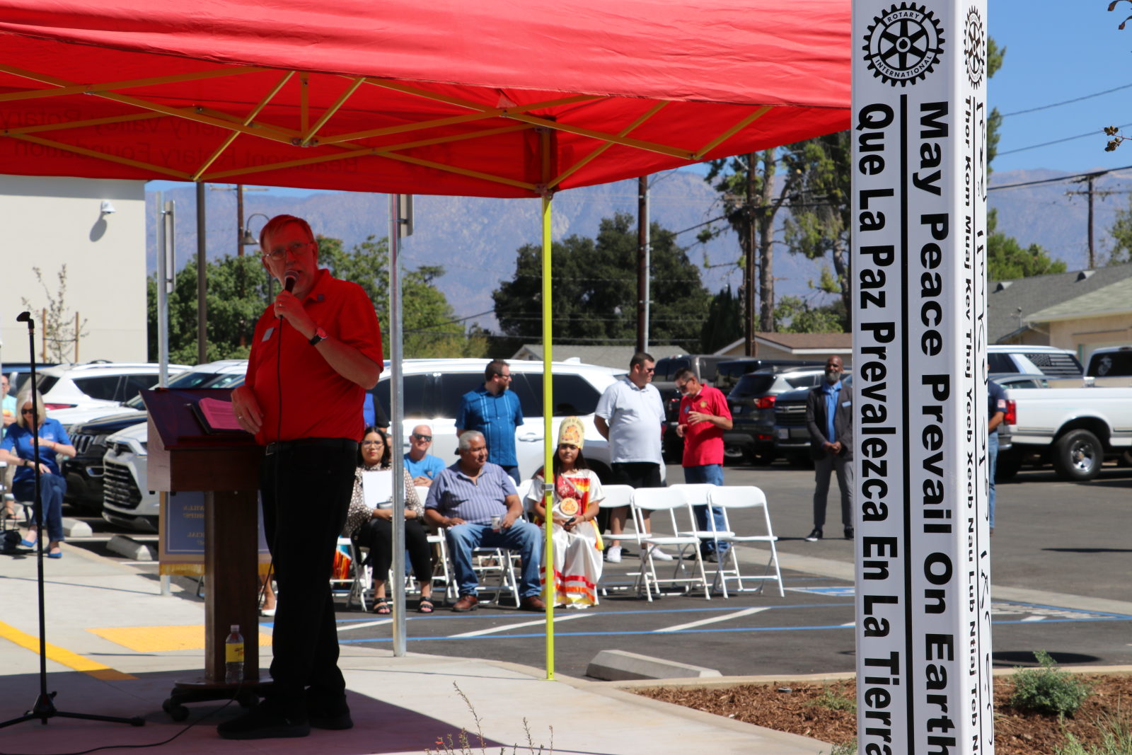 A Rotarian speaks at the podium under a red canopy beside the new Peace Pole, with guests seated in the library parking lot and mountains behind